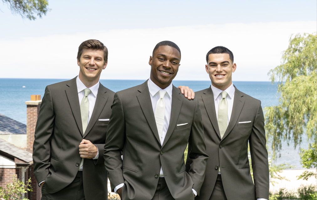 Three men in matching dark suits with light green ties stand outdoors, smiling, with a scenic view of trees, rooftops, and a large body of water in the background. One man has his hand on another’s shoulder. Should I rent or buy my suit or tuxedo?